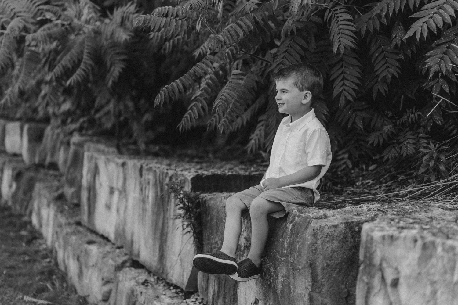 A child sits on a stone wall with ferns in the background in black and white.