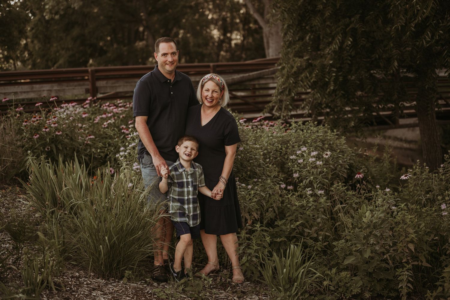Family of three posed in garden setting during golden hour with rustic wooden fence.