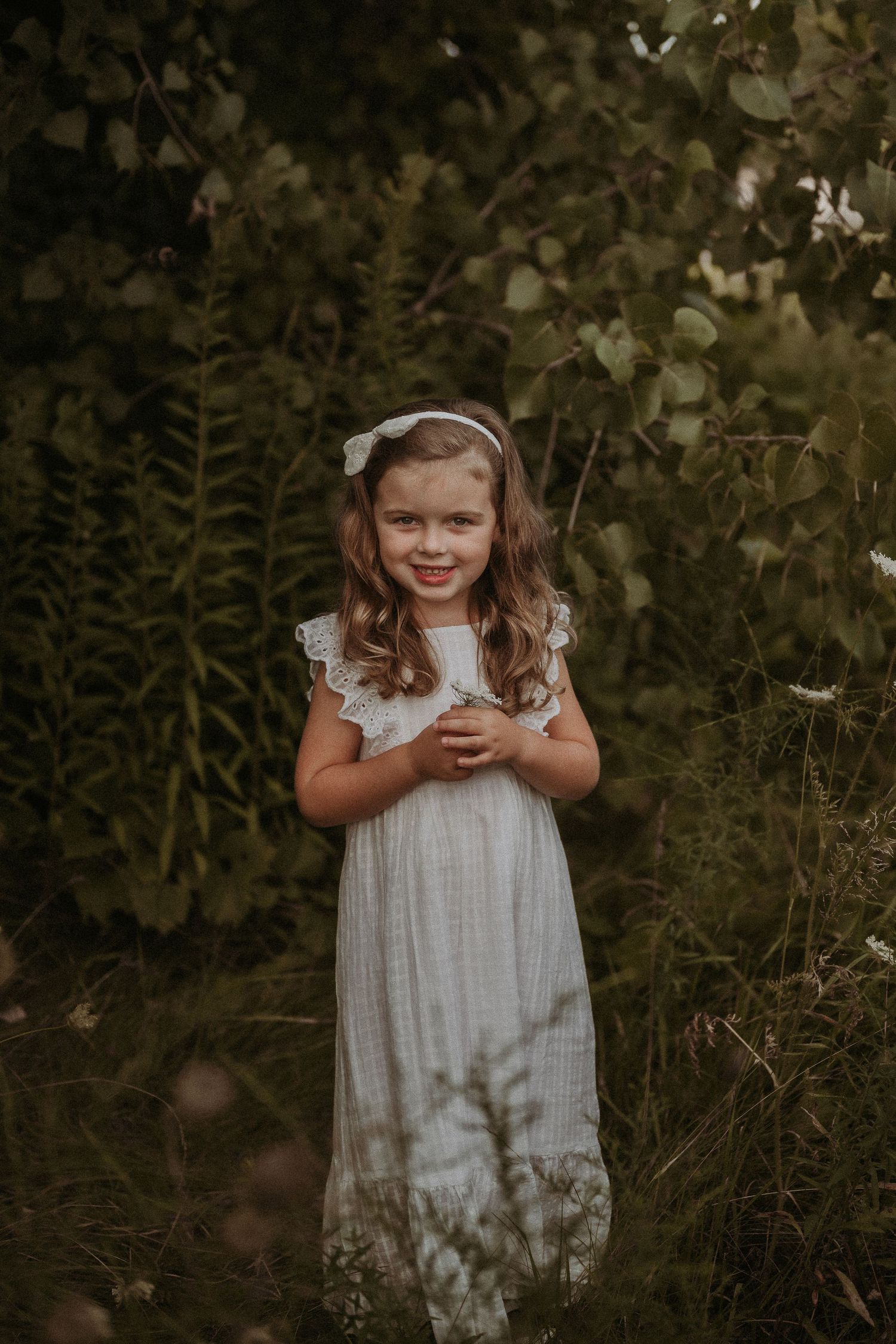 A child in a white dress stands in a natural setting surrounded by green foliage.