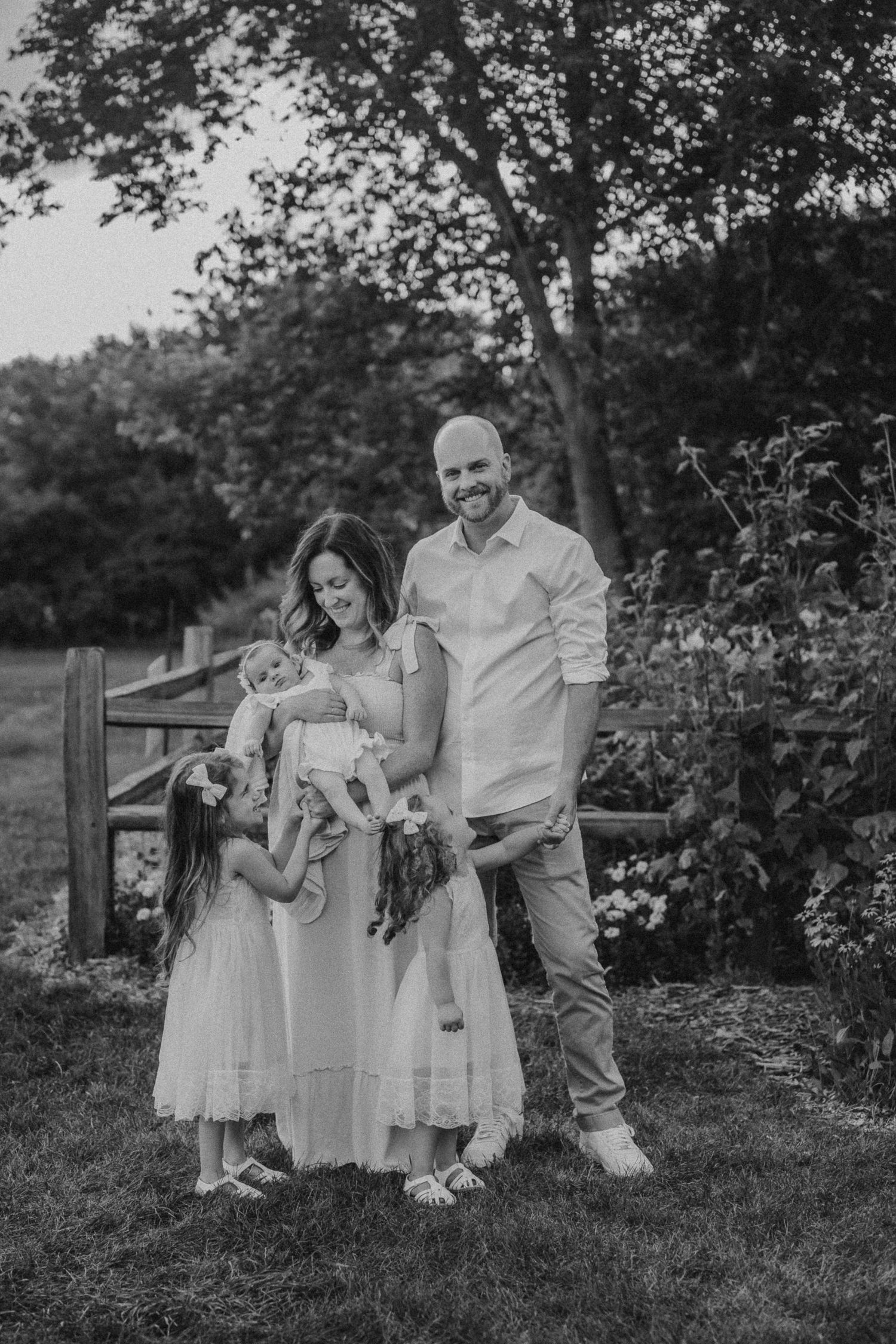 Black and white family portrait showing tender moments in a garden setting with rustic fence.