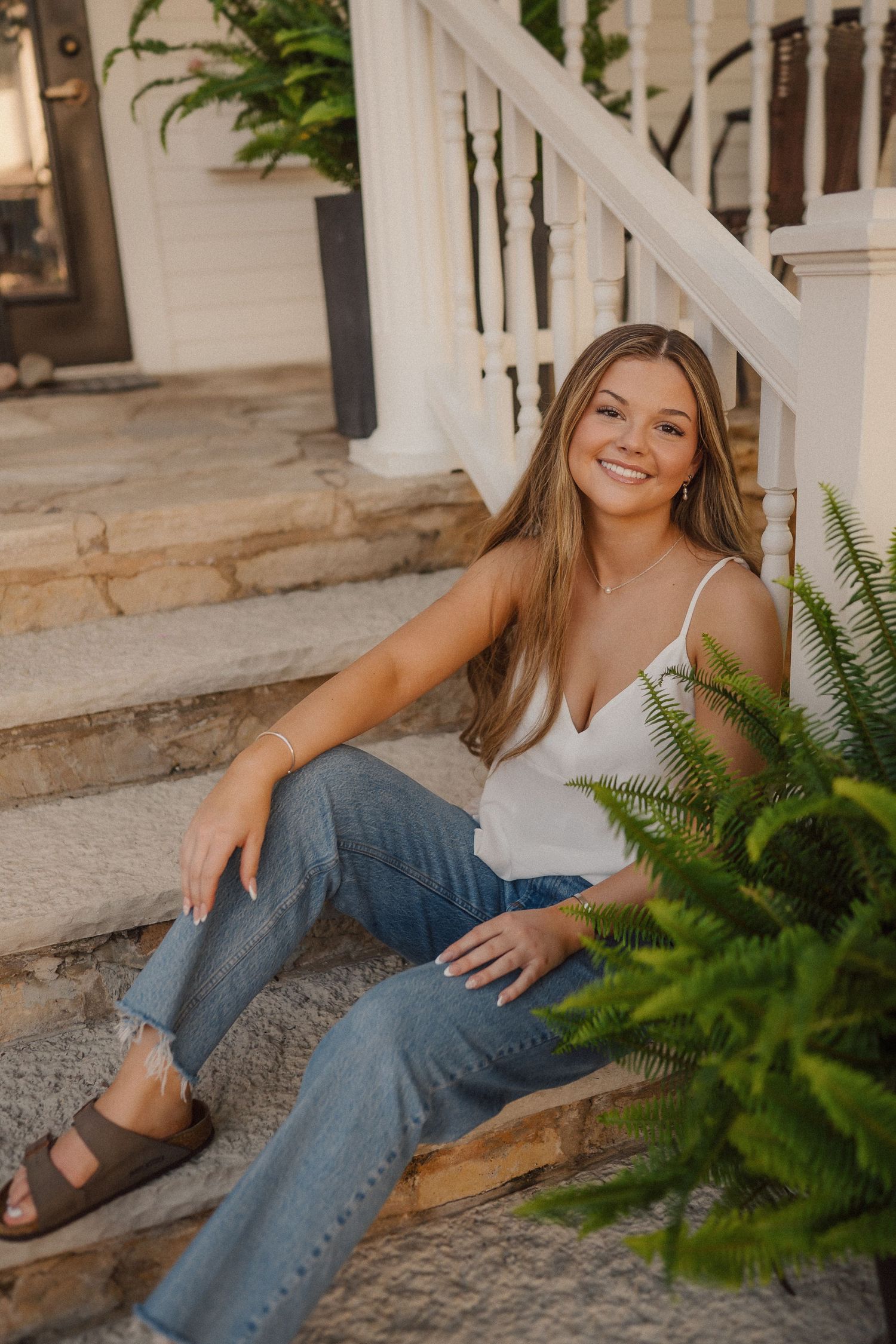 A woman relaxes on white porch steps surrounded by decorative ferns and plants.