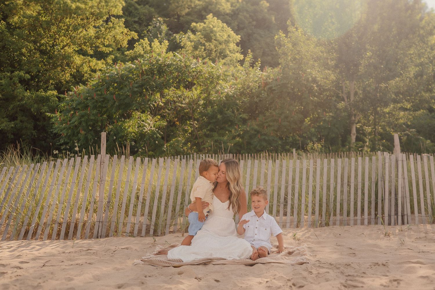 People in white outfits sitting together on sandy beach near wooden fence at sunset with trees in background.