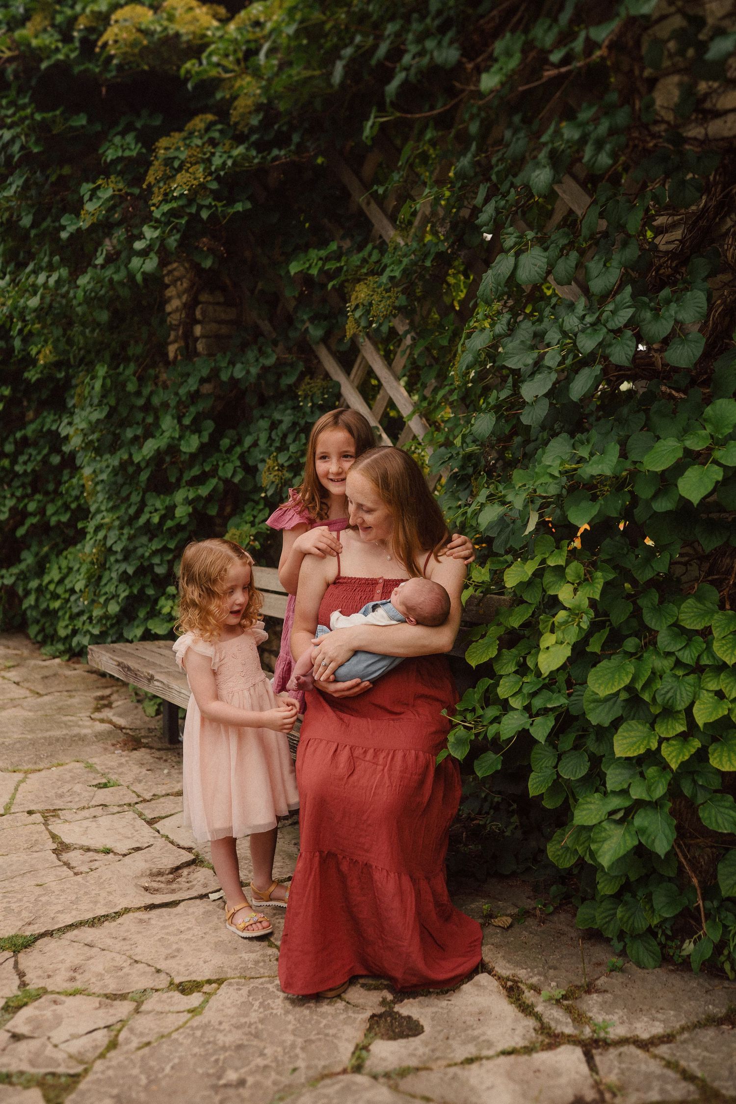 A tender family moment captured in a garden setting with lush green vines, featuring red and pink clothing on a stone path.