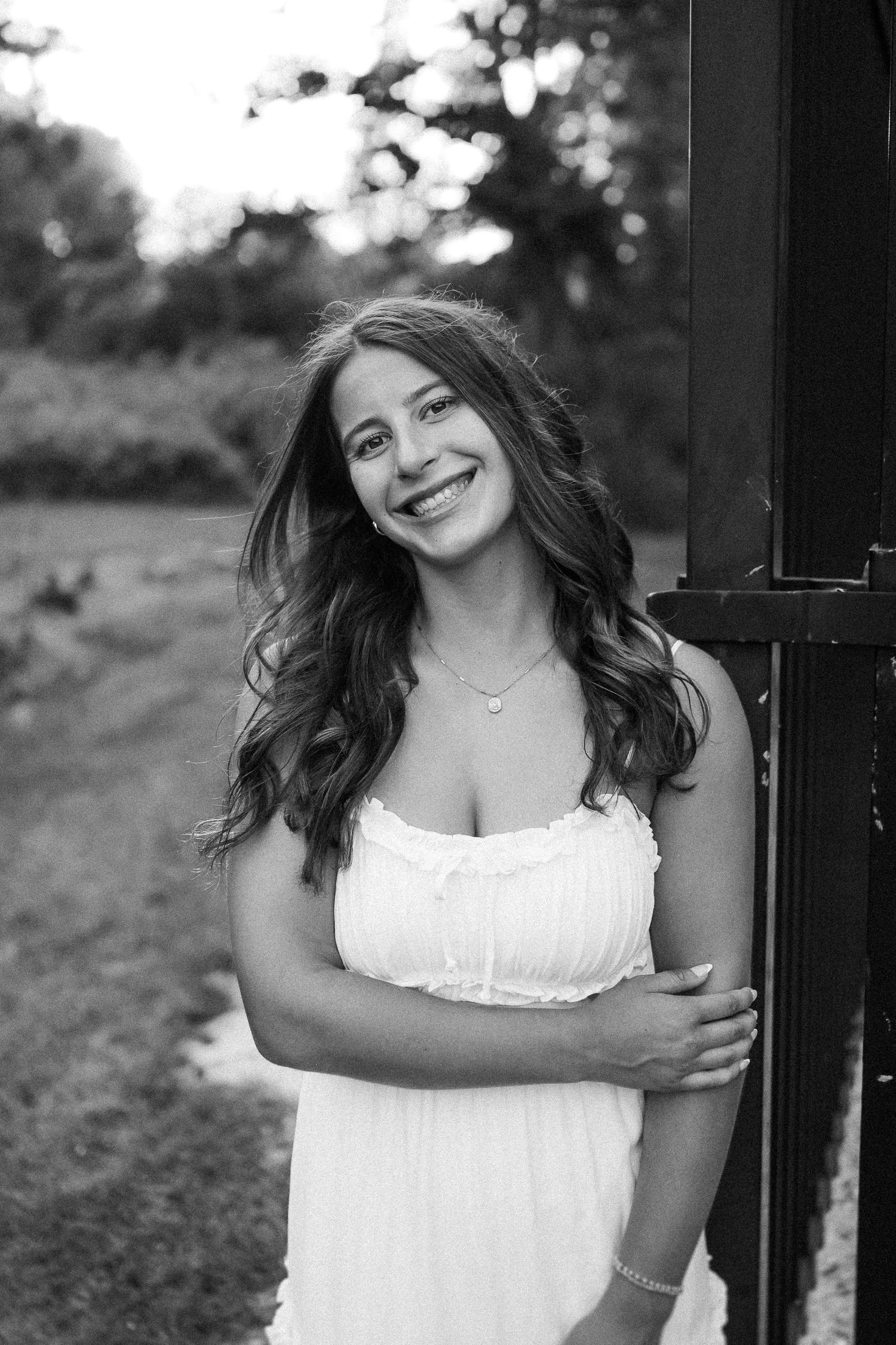 A person in a white sundress stands by a dark post outdoors in black and white photography.