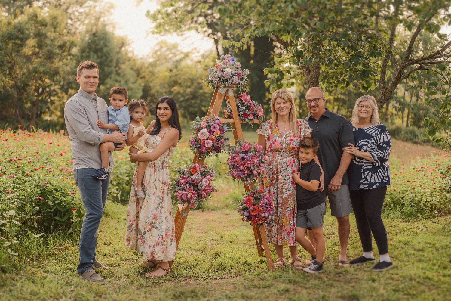 A group gathers outdoors for photos under trees with pink floral decorations and wooden ladders.