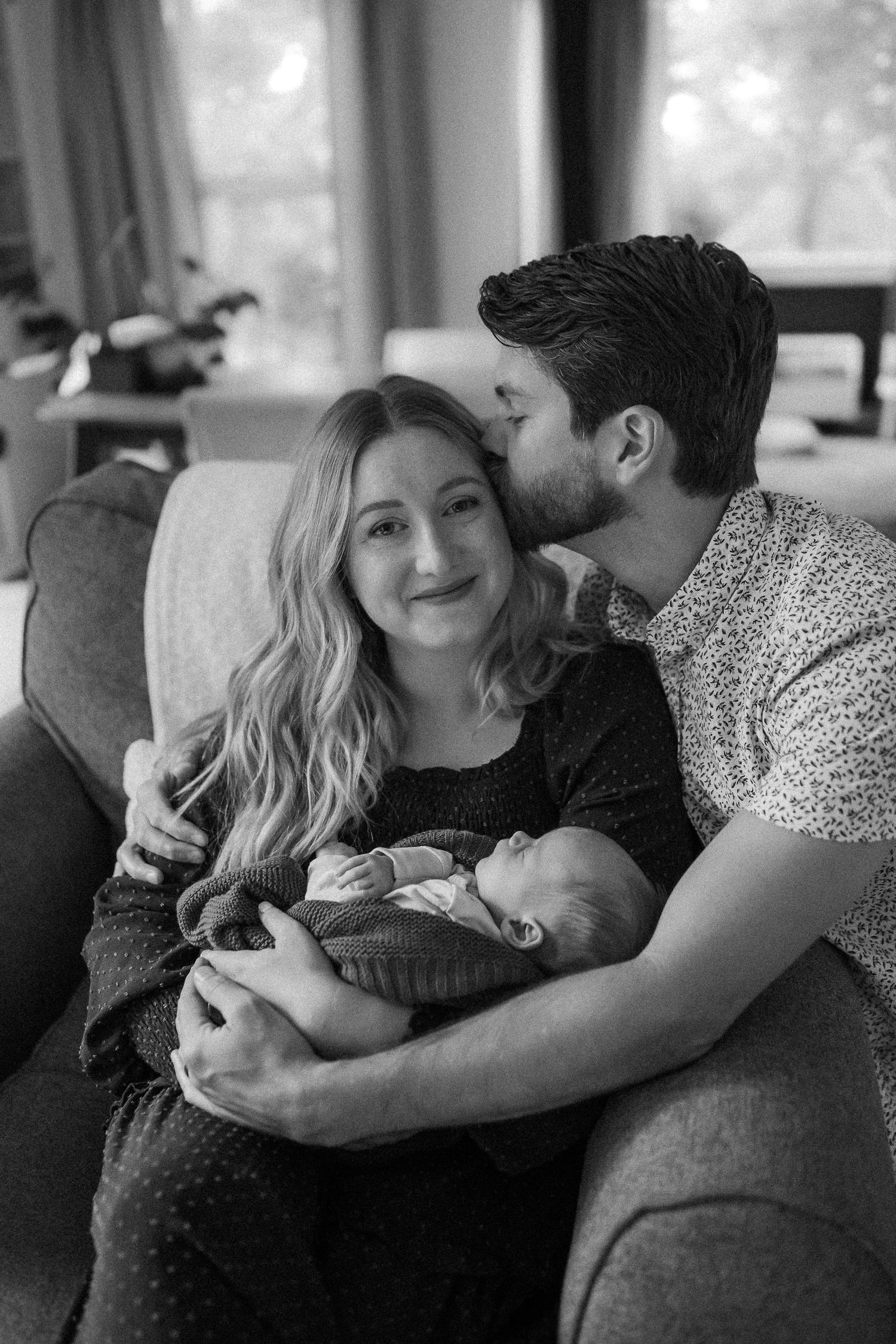 A tender black and white moment between parents cuddling their newborn baby on a cozy living room couch.