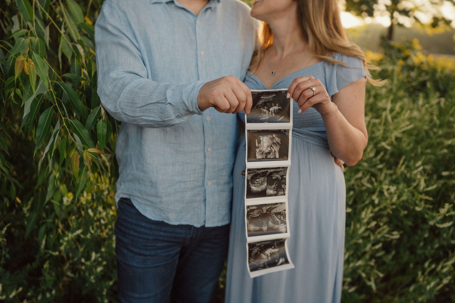 A couple in blue and denim clothing holds a strip of ultrasound photos outdoors at sunset among green foliage.
