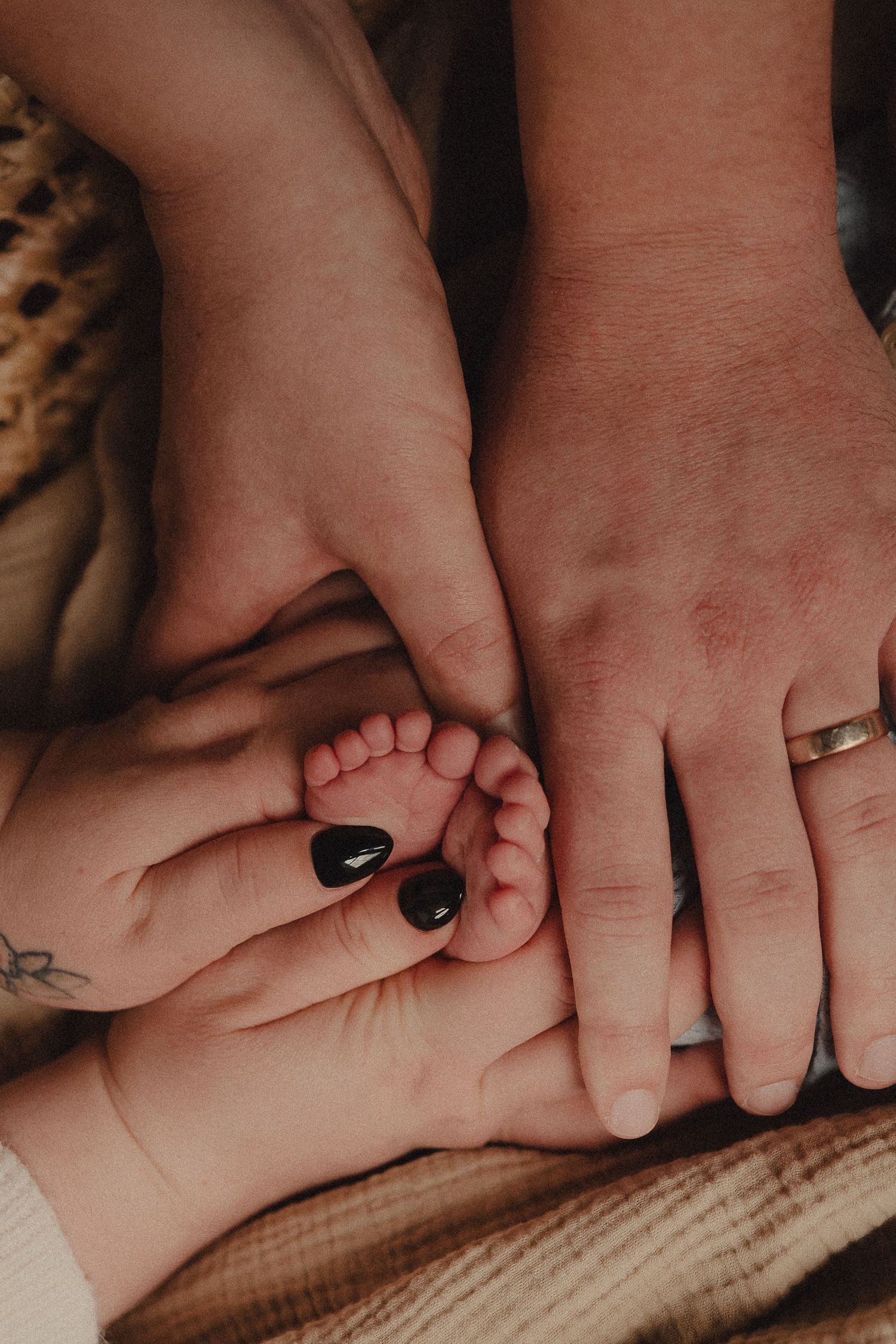 Parents cradling tiny newborn baby feet in their hands, mother with black nails and father wearing a gold wedding ring.