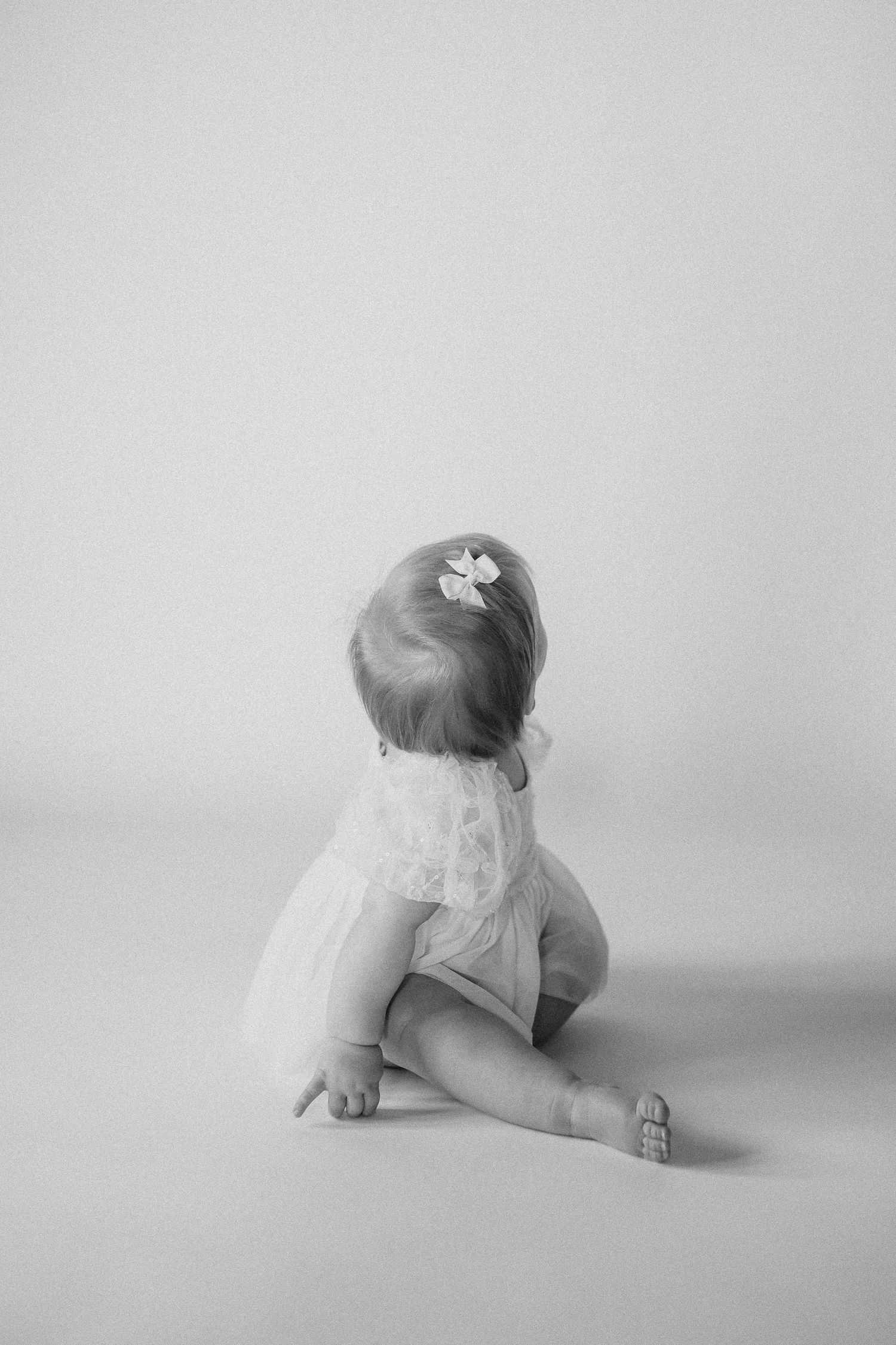 Black and white photo of a baby girl in a lace dress sitting with her back to the camera with a bow.