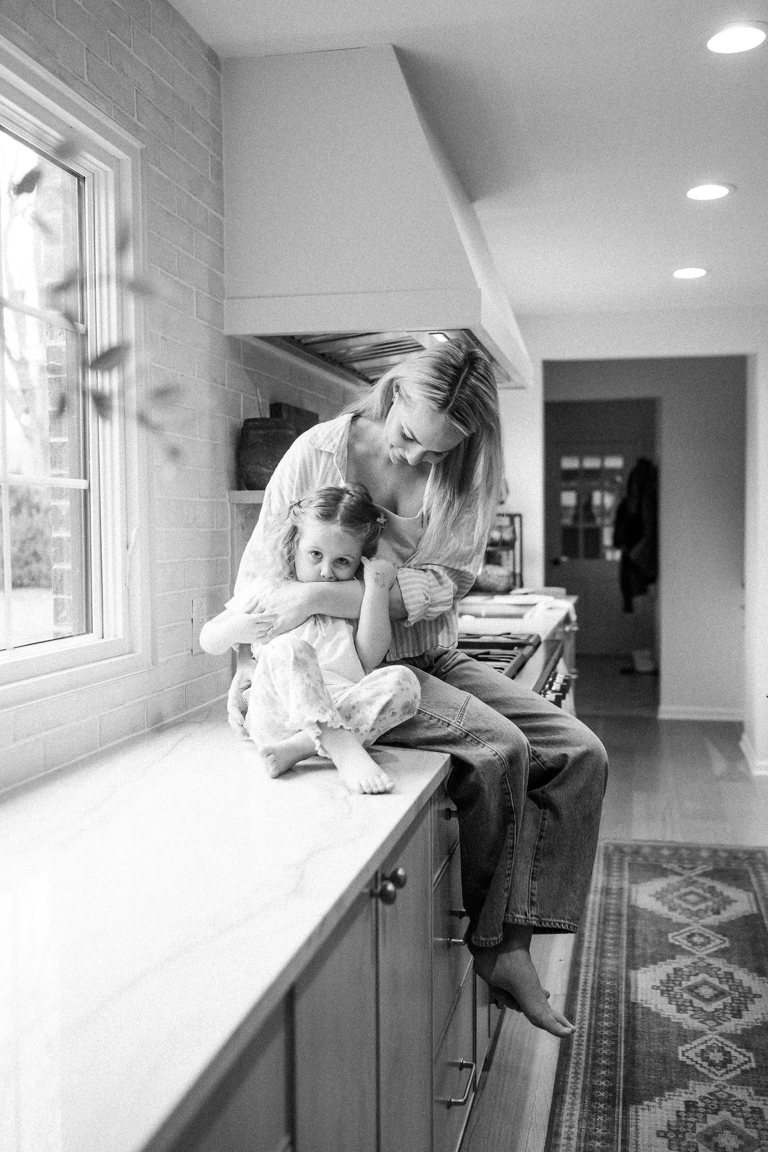 Black and white photo of a mother hugging her young daughter while sitting on a kitchen counter by a window.