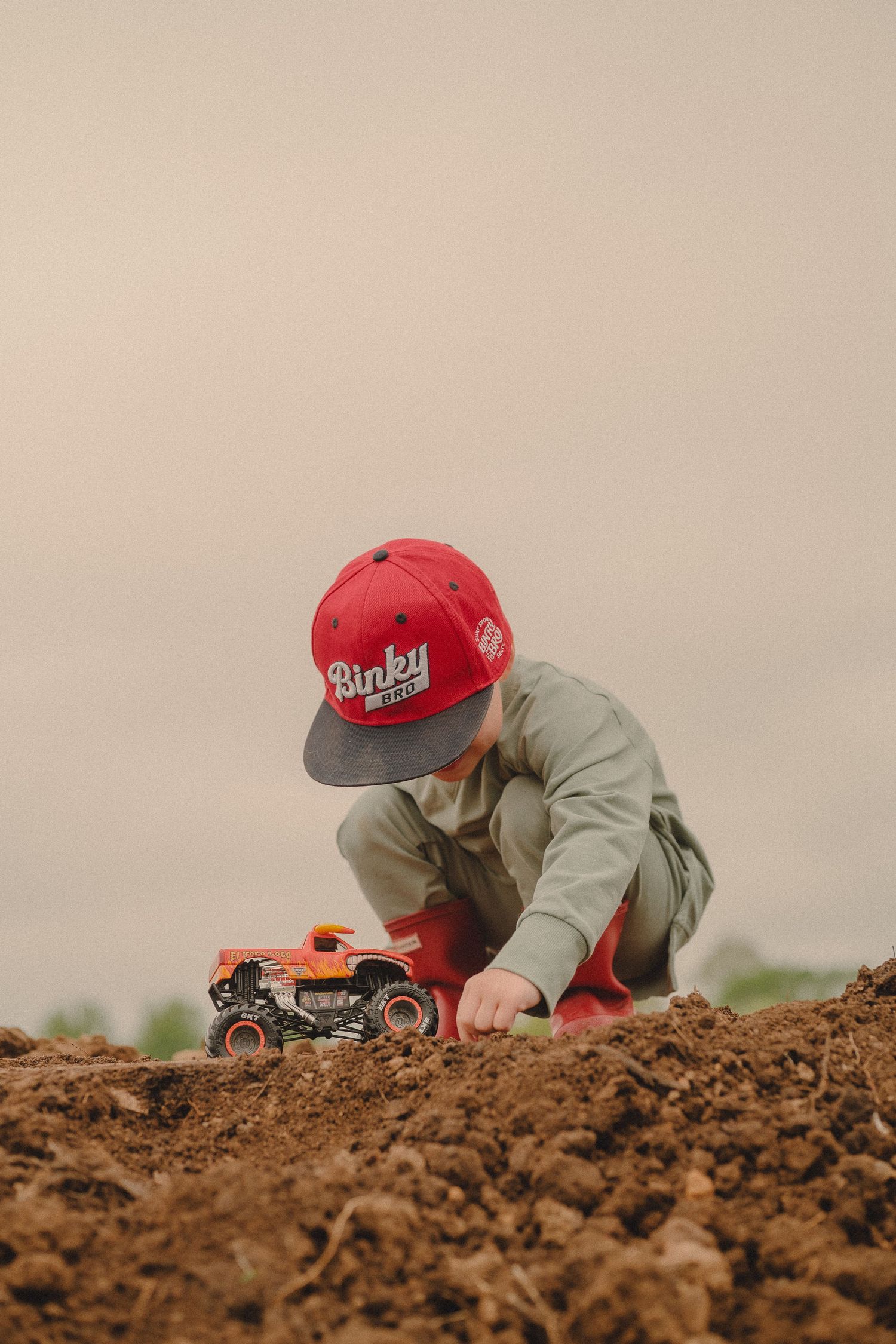 Young boy in red cap plays with toy monster truck in dirt field outdoors.