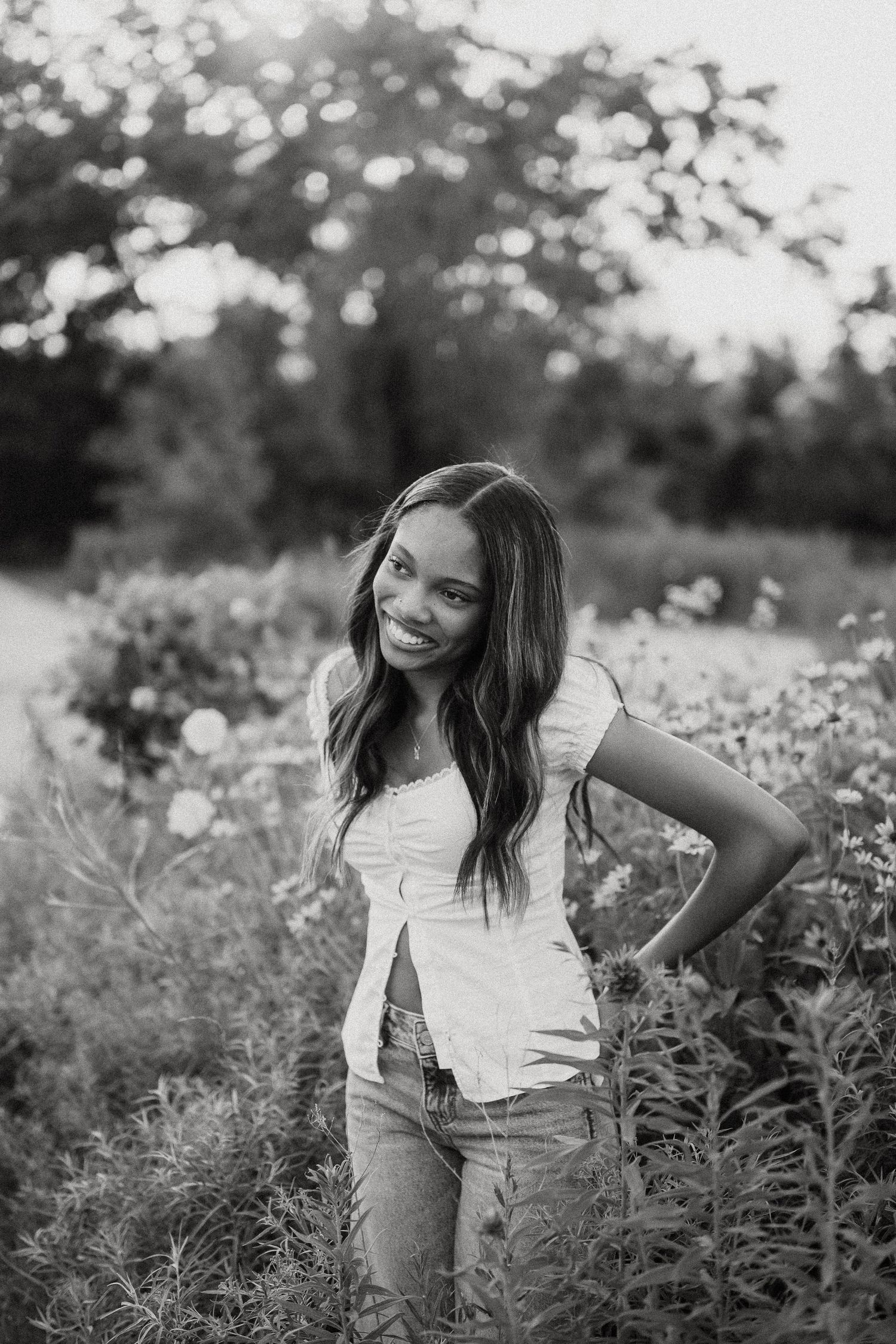 Black and white portrait of a smiling young woman in a white top and jeans posing in a lush flower field.