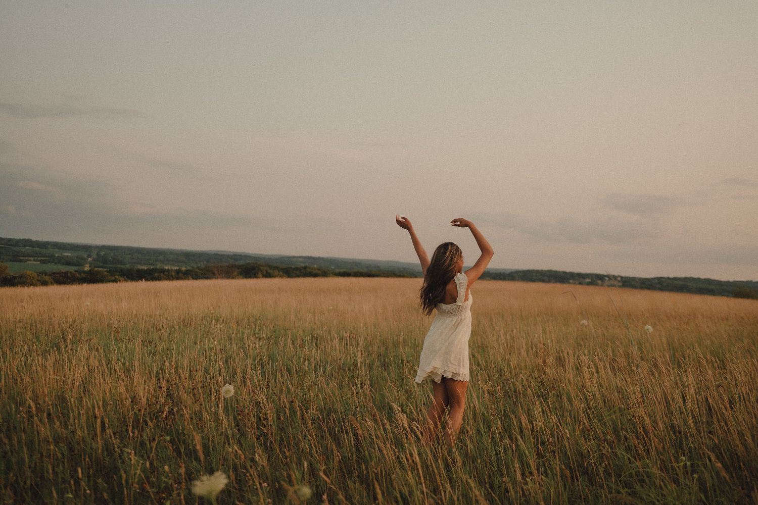 Young woman in white dress dancing freely in golden wheat field at sunset with rolling hills in background.