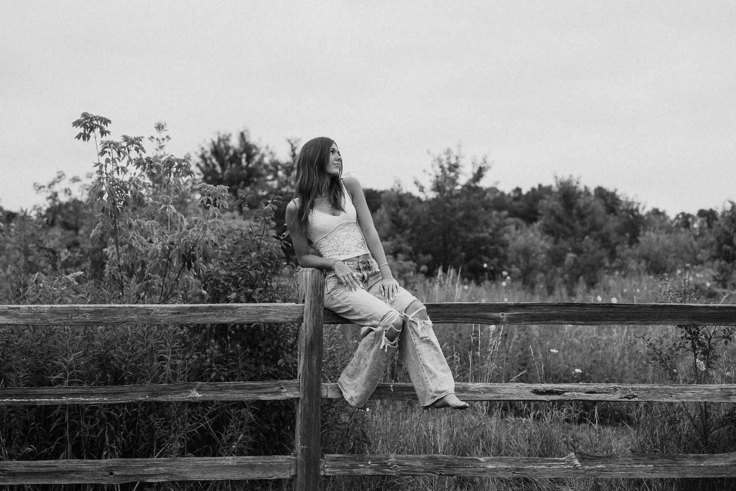 Young woman in white tank and wide-leg jeans leaning on rustic wooden fence in rural landscape, black and white photo.