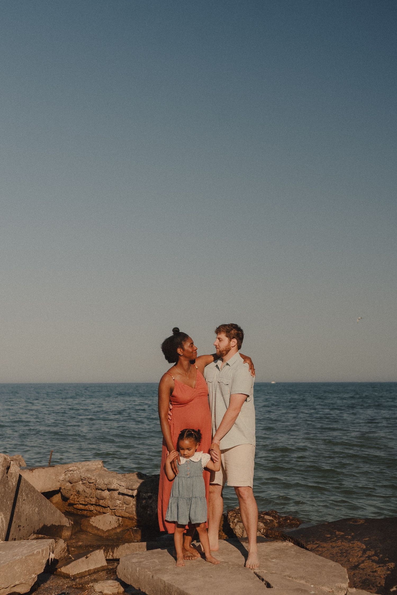 An interracial couple with their toddler daughter pose on rocky shoreline under a clear blue sky.