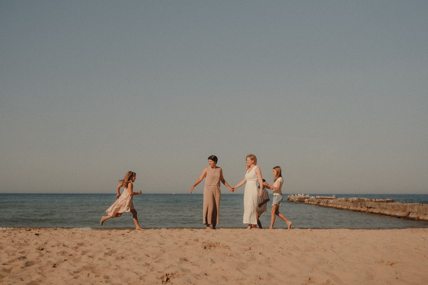A family of four walks hand-in-hand along a sunny sandy beach with calm ocean water in the background.