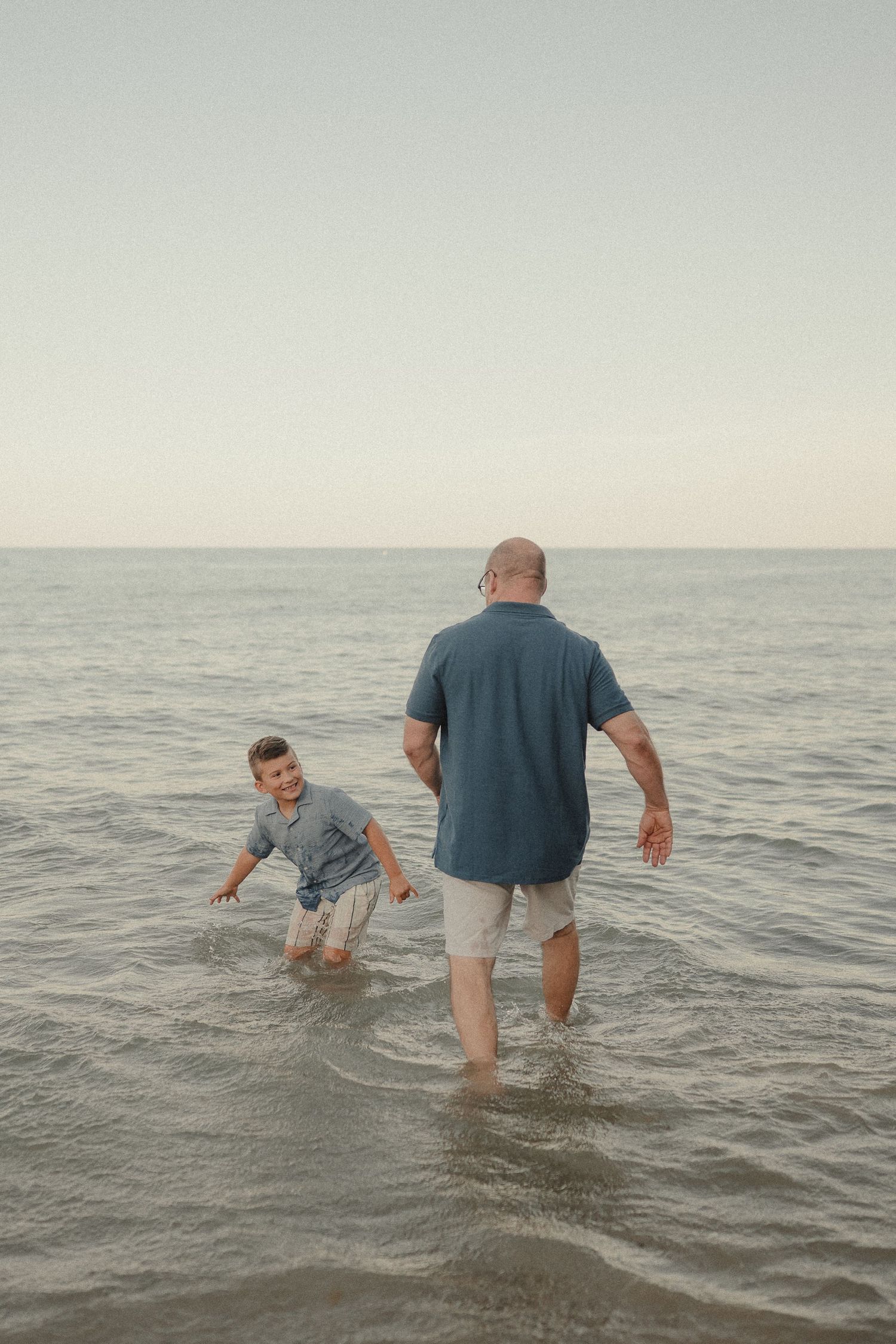 Father and son wading together in shallow ocean water at dusk, enjoying a playful beach moment.