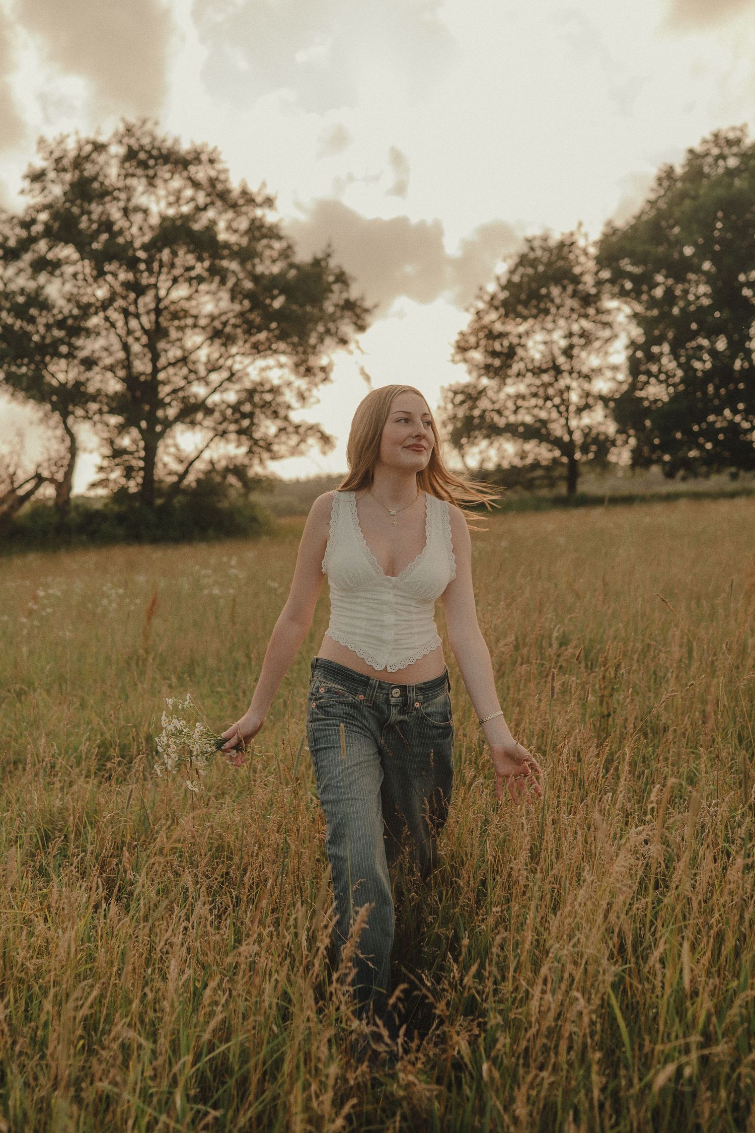 Young woman with red hair walks through golden field at sunset wearing white crop top and jeans.
