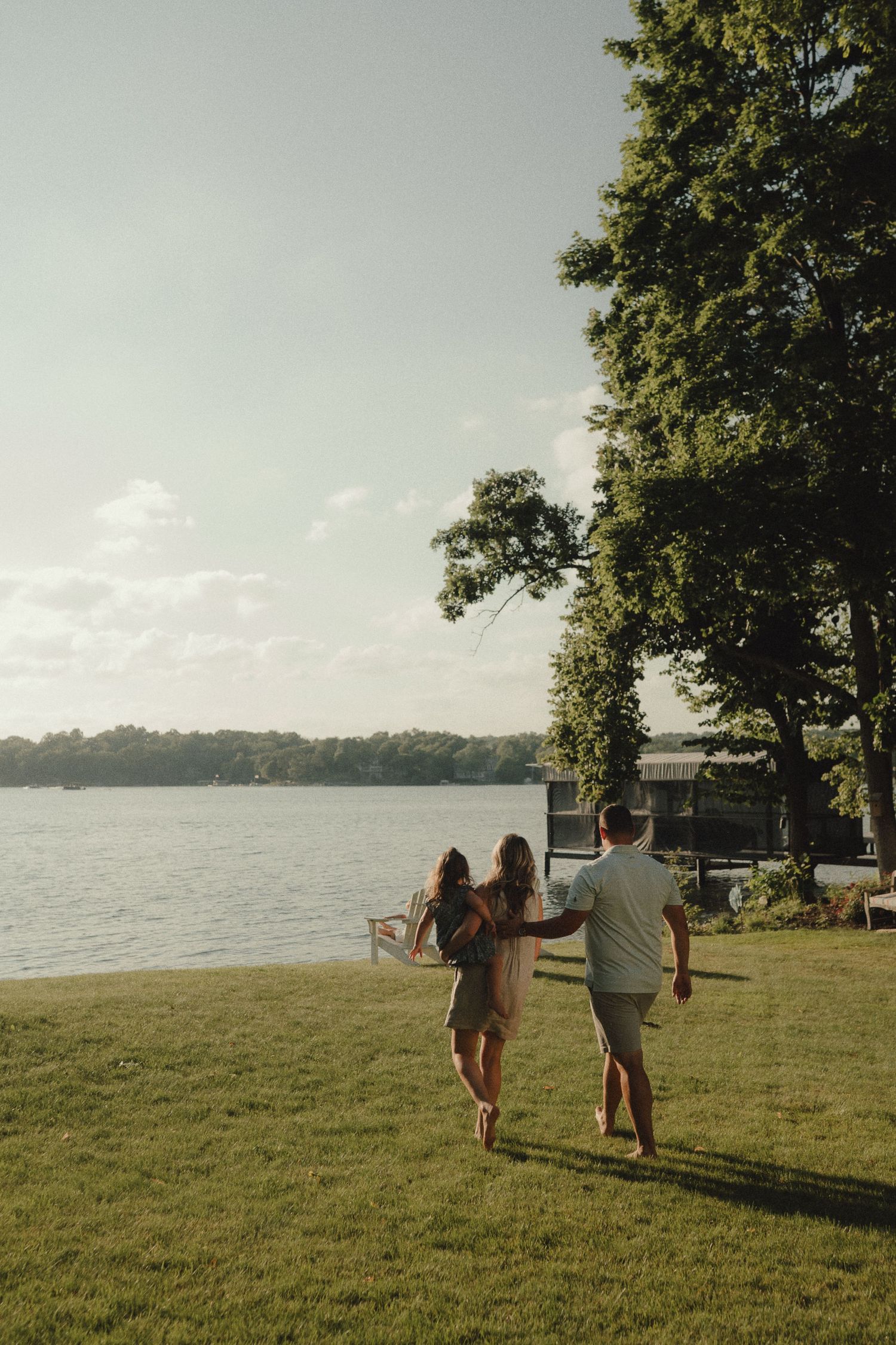 Couple walking hand-in-hand on green lawn toward serene lakeside at golden hour.