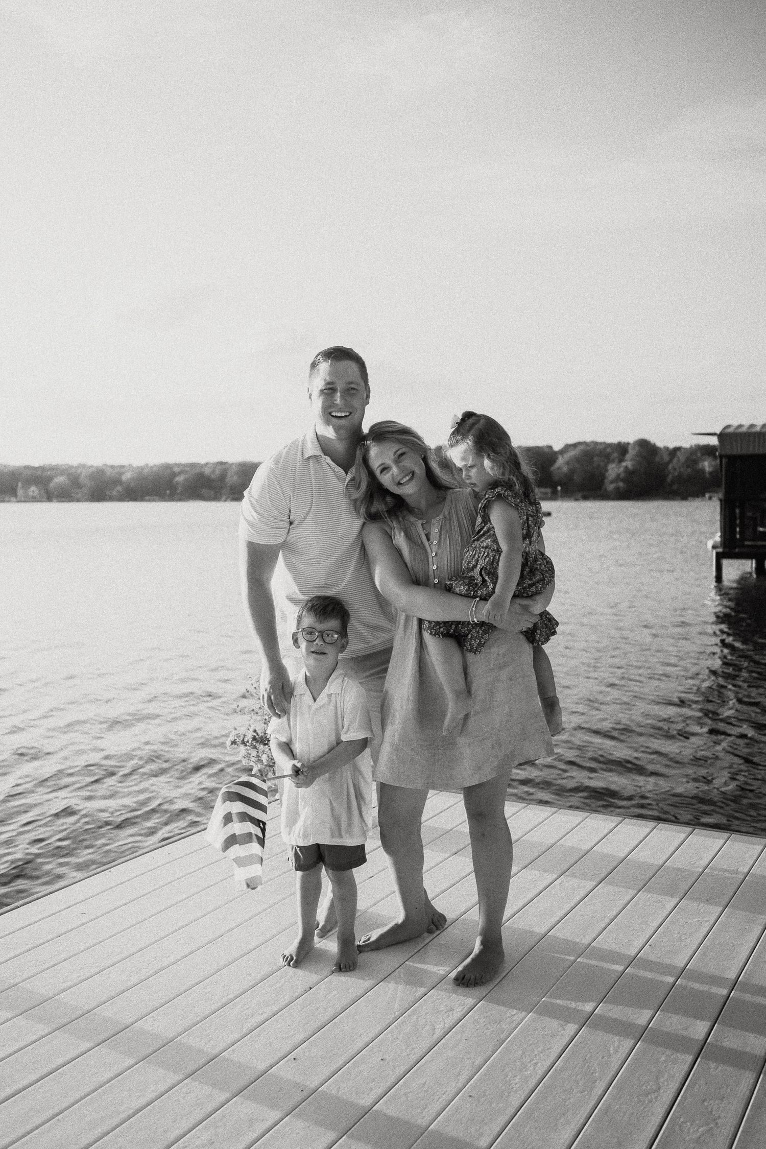 Black and white photo of a smiling family of four posing on a dock by the lake.