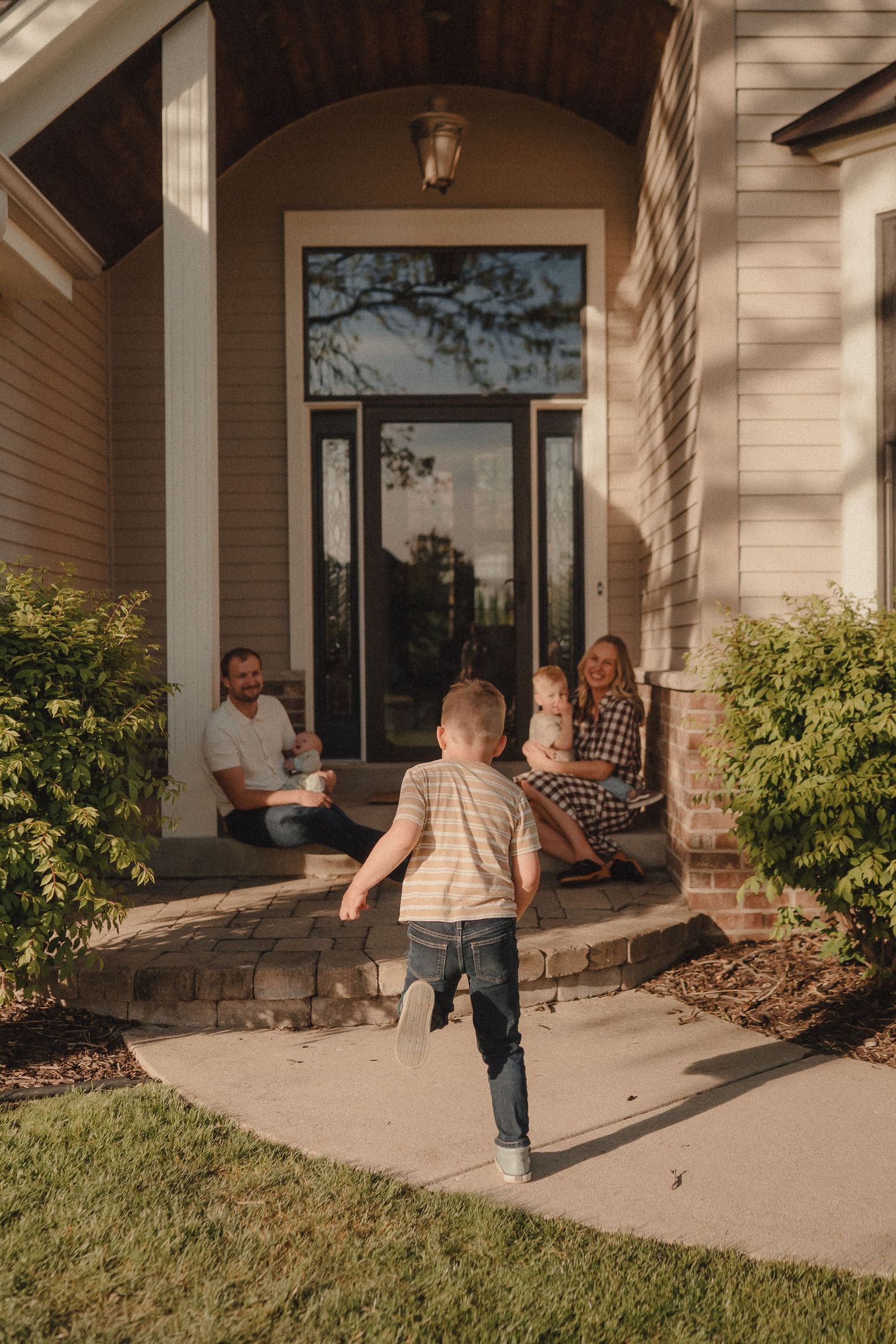 A young boy runs toward his family sitting on the front porch steps of a suburban home.