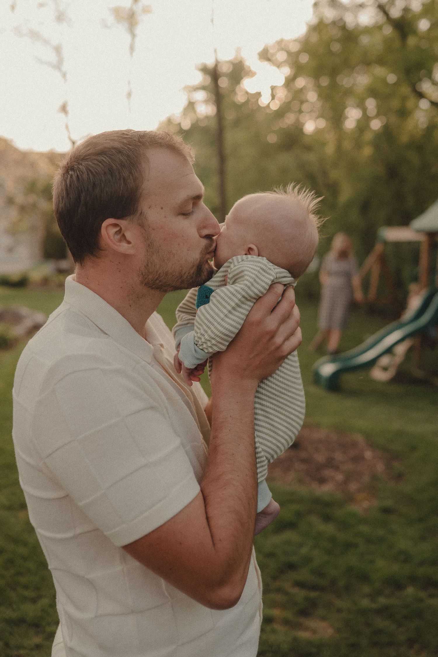 Loving father gently kisses newborn baby outdoors in backyard at golden hour sunset.