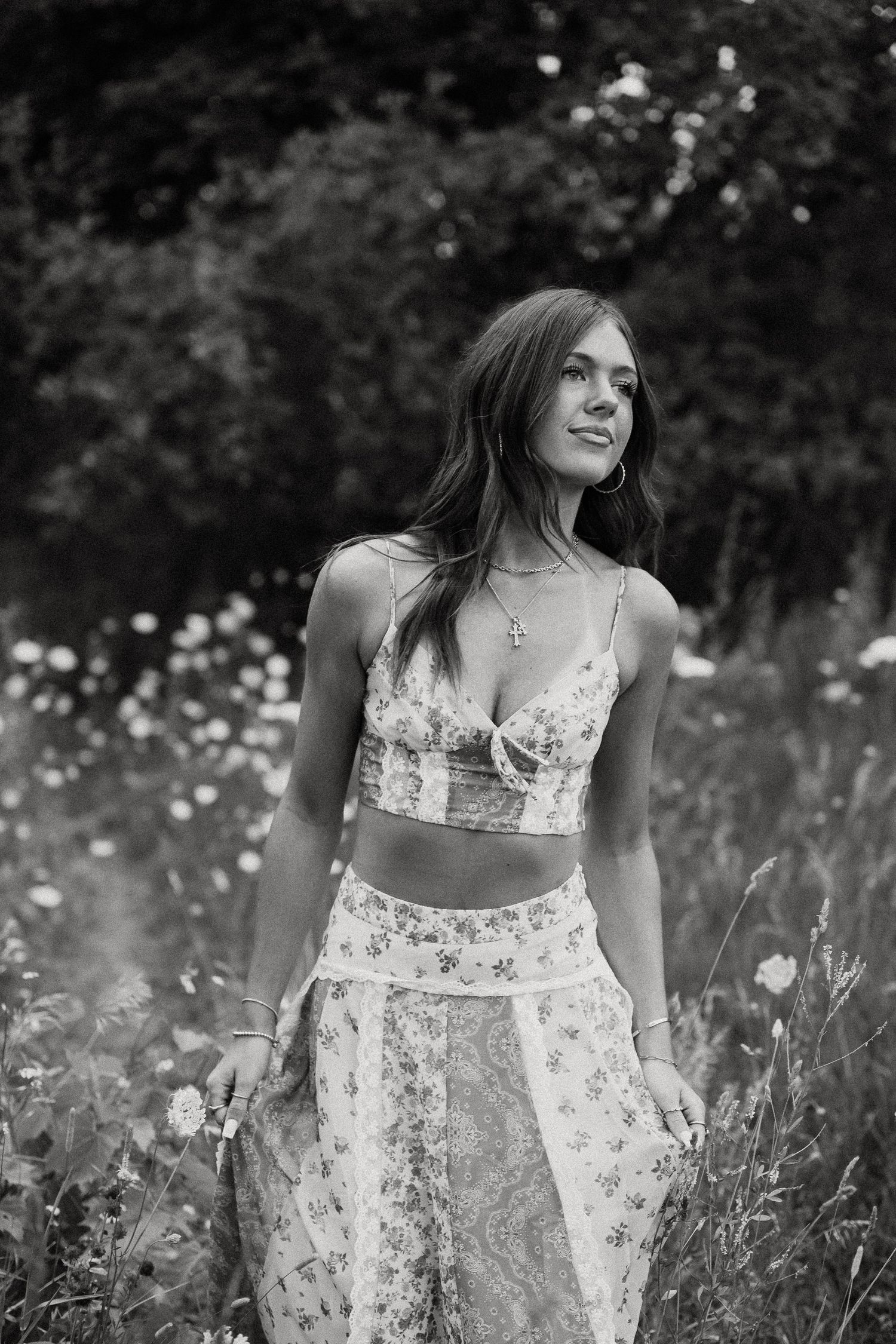 Black and white photo of young woman in floral crop top and skirt standing in wildflower field.