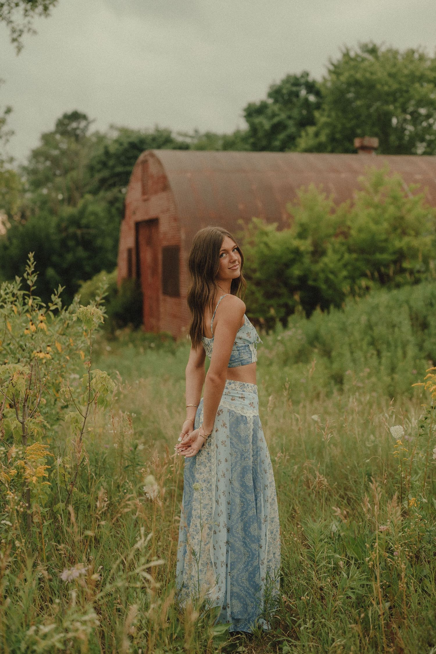 Young woman in blue floral two-piece maxi skirt set standing in wildflower field near rustic barn.