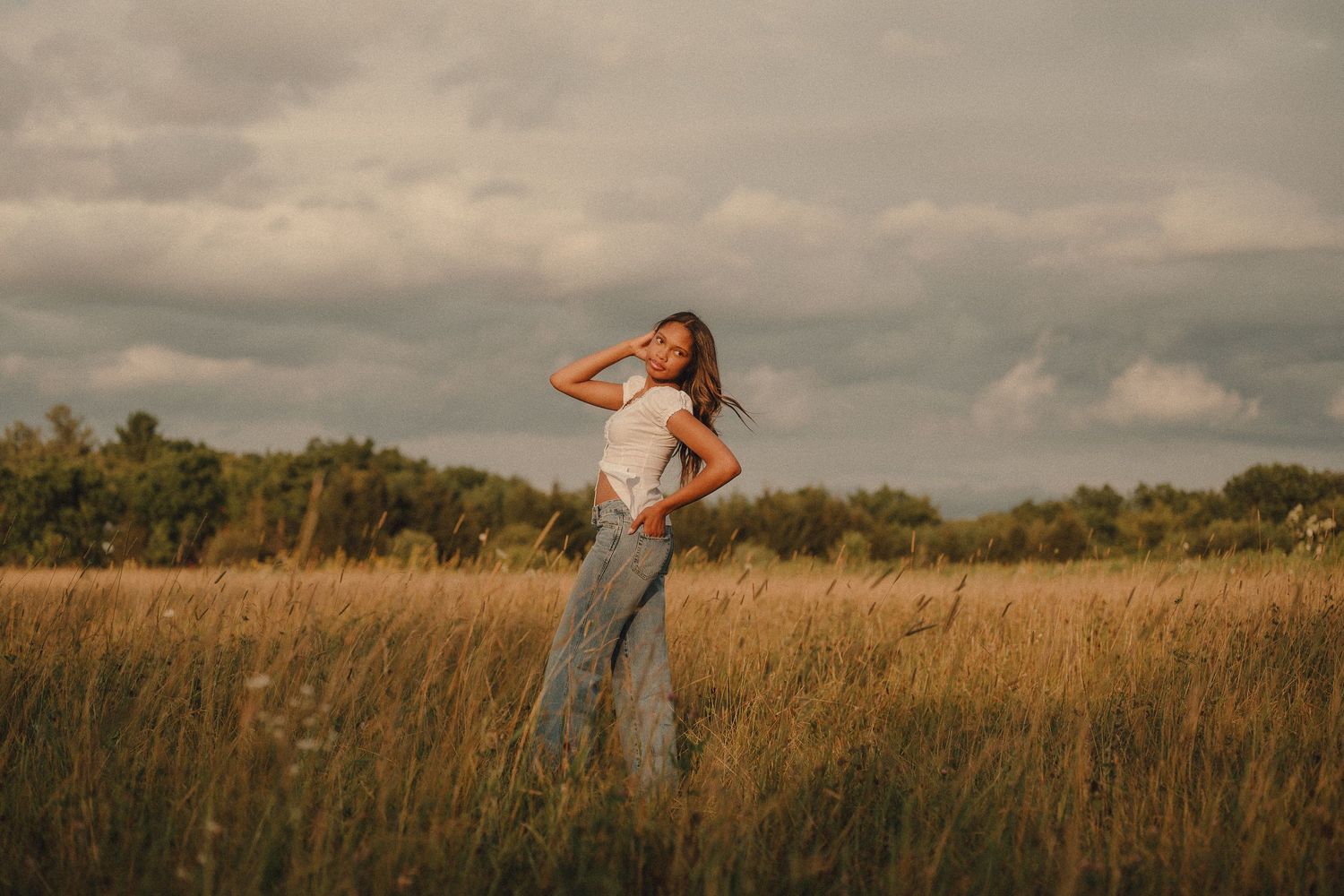 Young woman in white top and jeans standing in golden field under dramatic cloudy sky.
