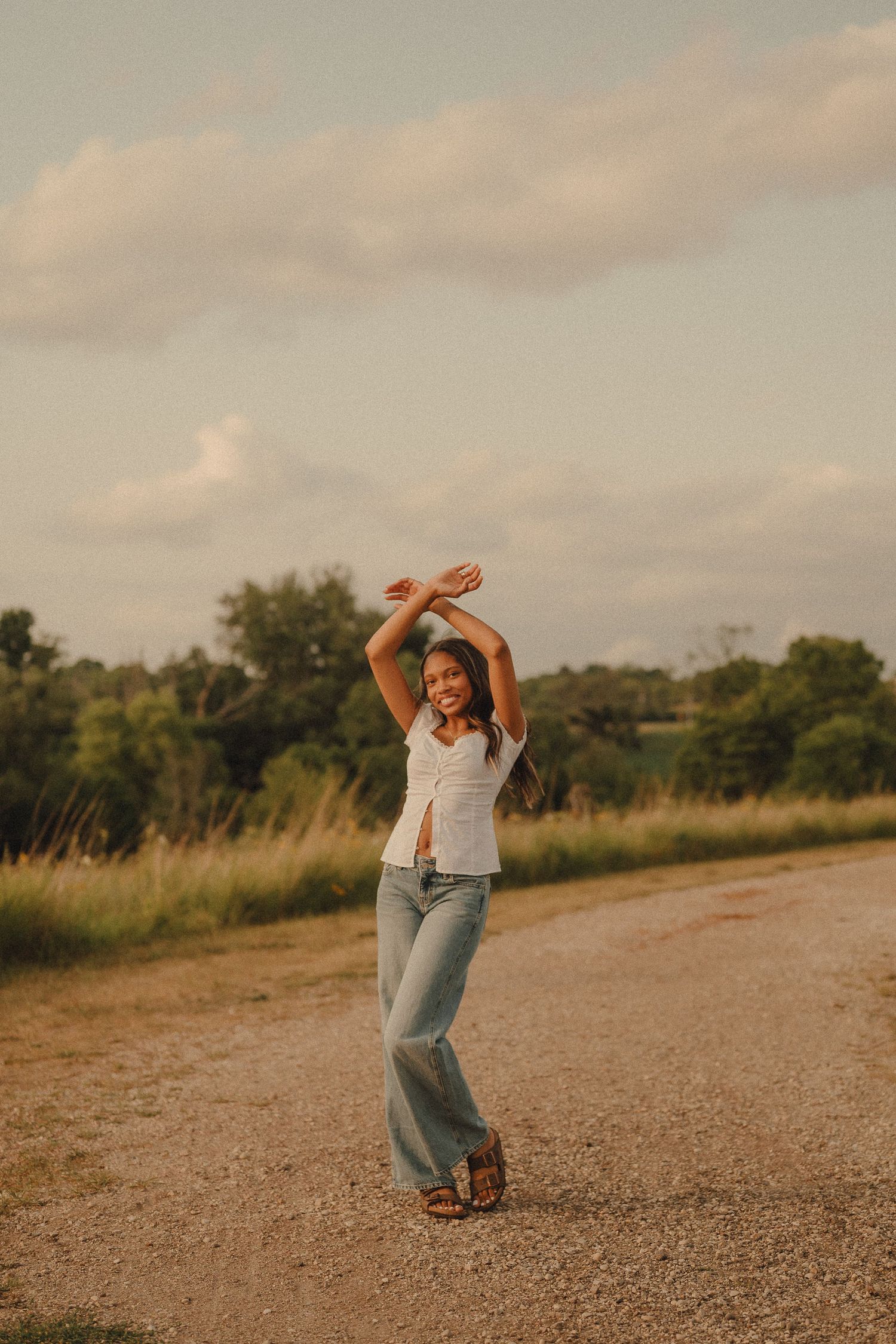 Joyful young woman dancing on a gravel path with arms raised, surrounded by golden meadow.