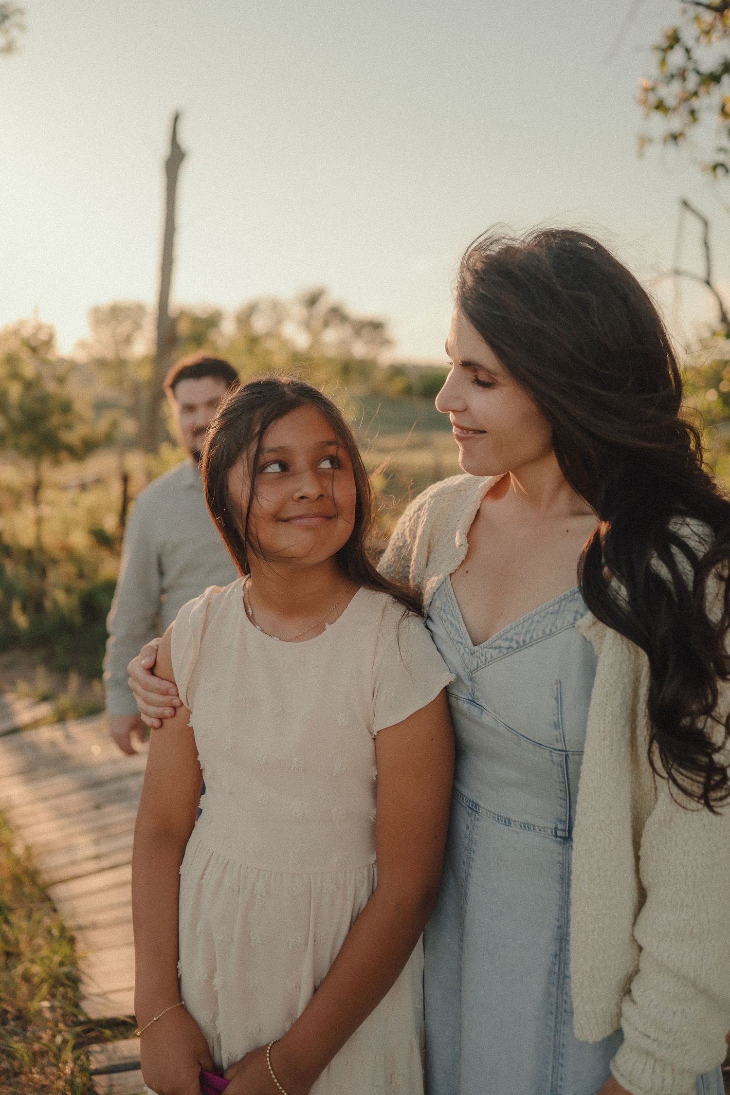 Woman in denim dress embraces young girl outdoors during golden hour family portrait.