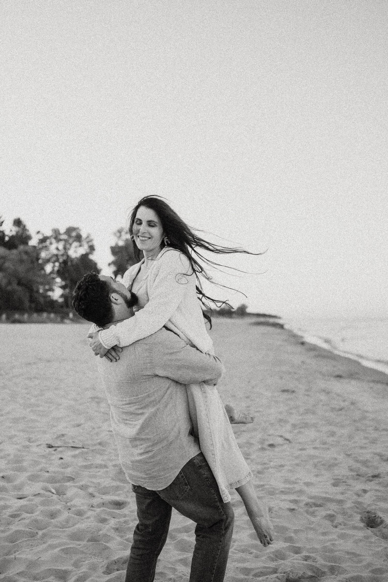 Black and white photo of a man lifting a smiling woman on a sandy beach with trees in the background.