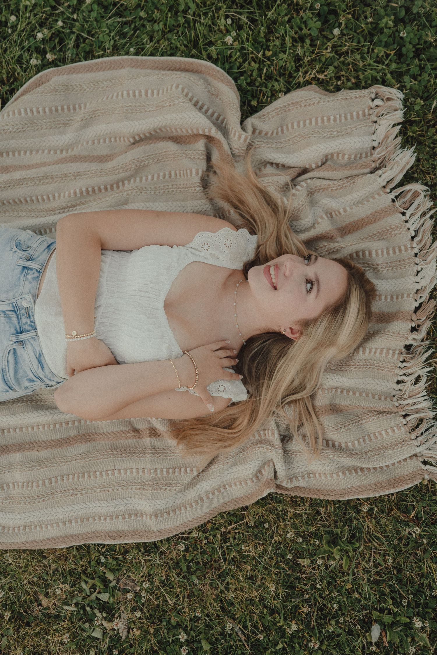 Young blonde woman lying on a striped fringe blanket on green grass, wearing white top and denim shorts.