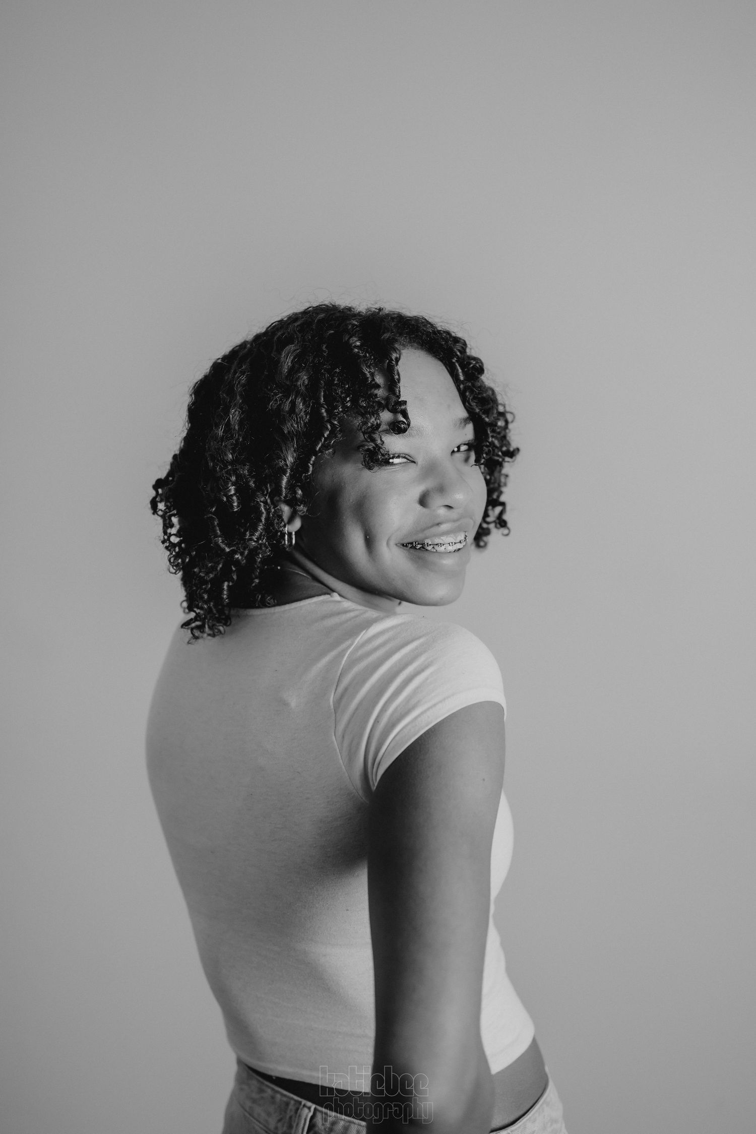 Black and white portrait of a person with curly hair smiling over their shoulder.