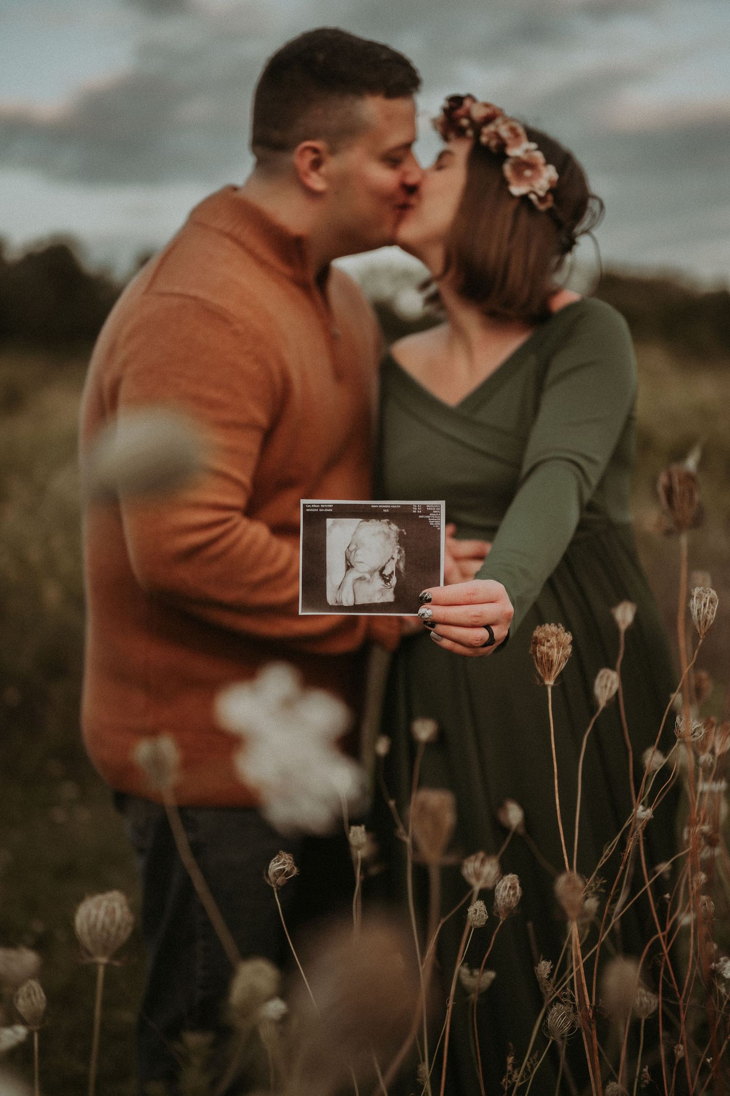 A couple holds an ultrasound photo while sharing a tender moment in a wildflower field.