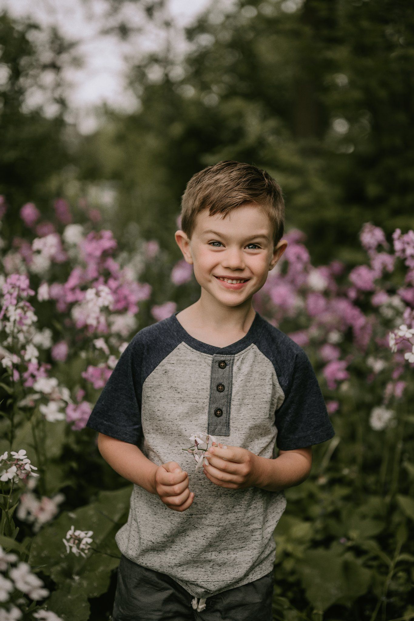 Child smiles while holding flowers in garden with pink blooms in background.