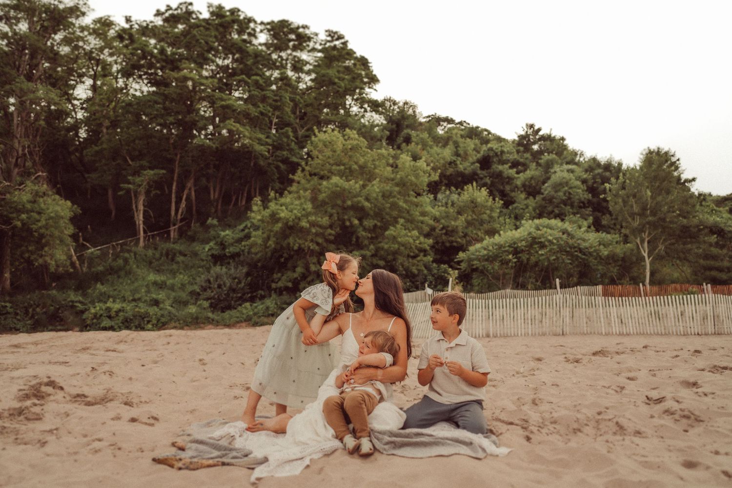 A family relaxes together on a blanket at the beach with trees in background.