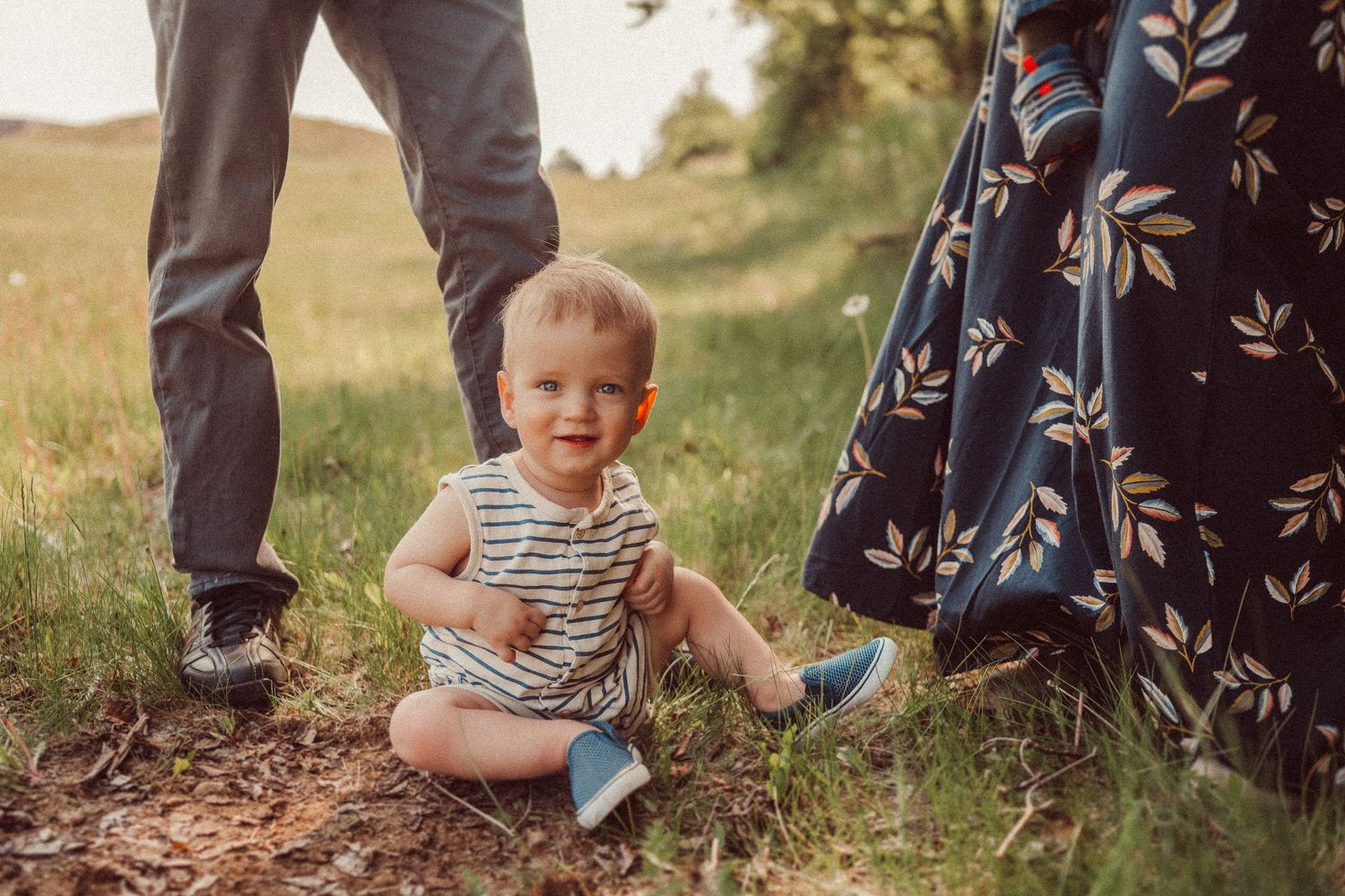 A toddler in striped clothing sits on a path while adults stand nearby.