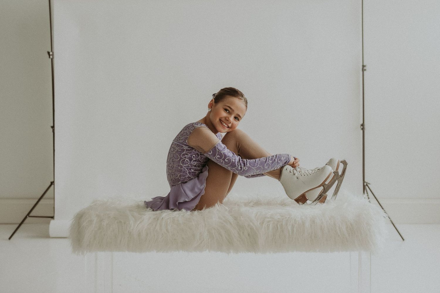 Ice skater adjusts skate while posing on white fur surface in studio setting.