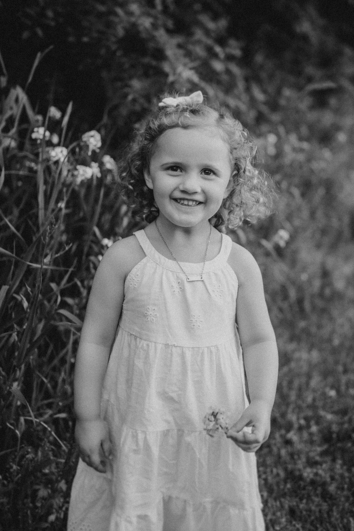 A sweet black and white portrait of a child in a white sundress standing in a garden with wildflowers.