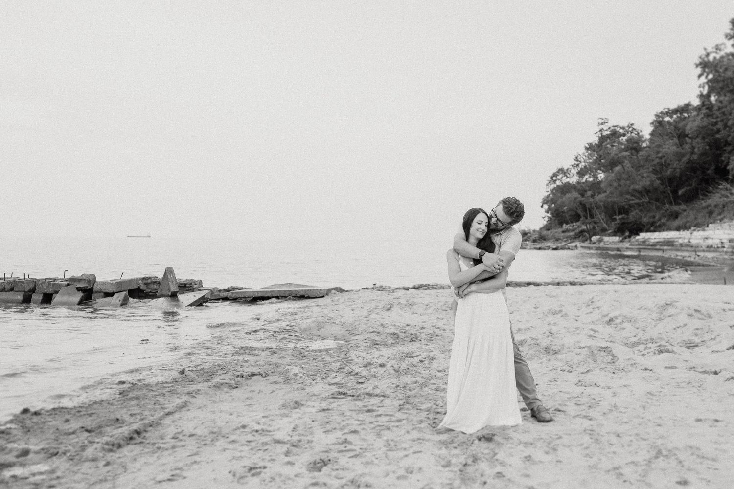 A couple embraces on a rocky beach during a black and white photoshoot with ocean waves in the background.