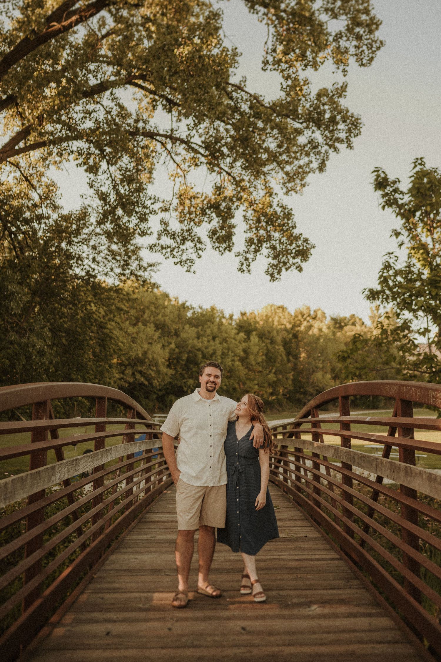Couple standing together on wooden bridge during sunset with autumn trees overhead.