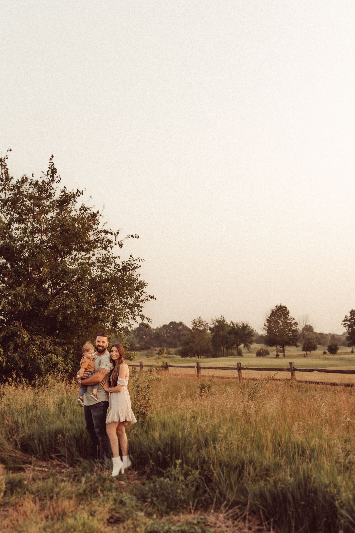 Sunset family portrait in a pastoral setting with wooden fence and trees creating a natural backdrop.
