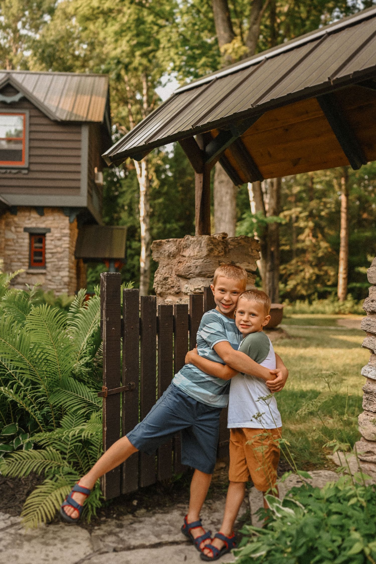 Two young boys hugging and laughing by wooden gate of rustic cabin.