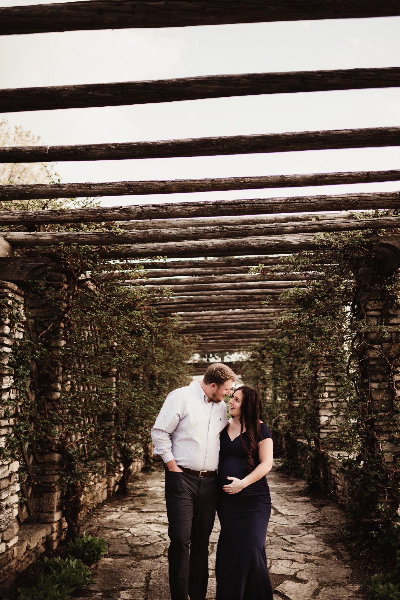 Couple shares intimate moment under wooden pergola during maternity photoshoot.