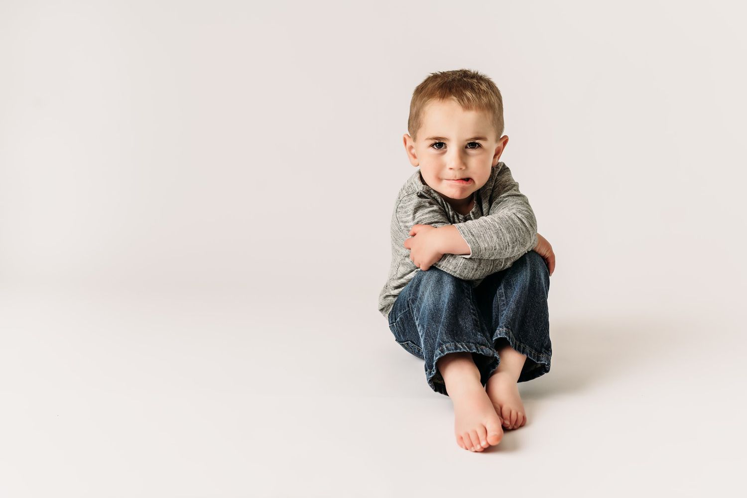 Child in gray sweater sits cross-legged on white floor during photo session.