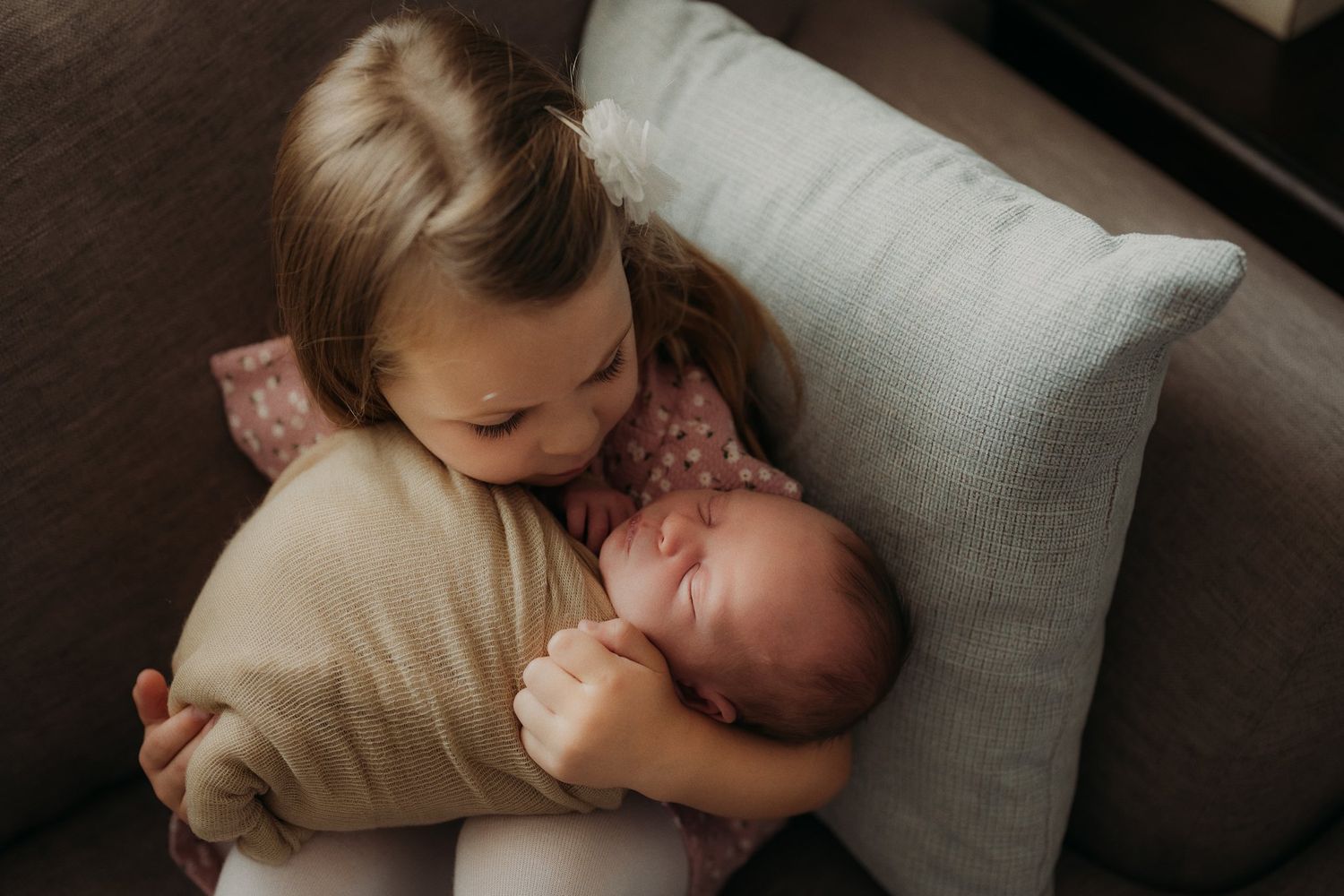 Intimate indoor portrait of a child cradling a newborn baby on a couch.