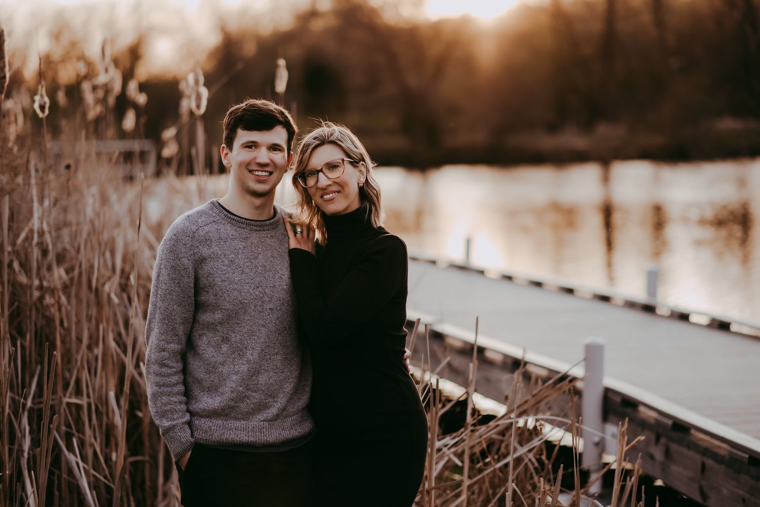Couple poses together near waterfront during golden hour with cattails in background.