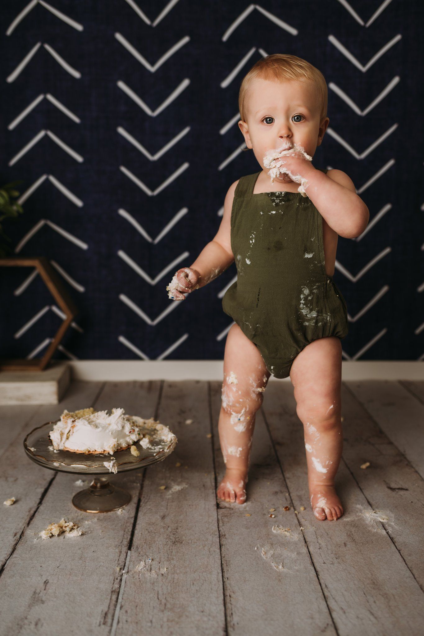 Standing toddler in olive romper covered in cake crumbs on wooden floor with demolished dessert.