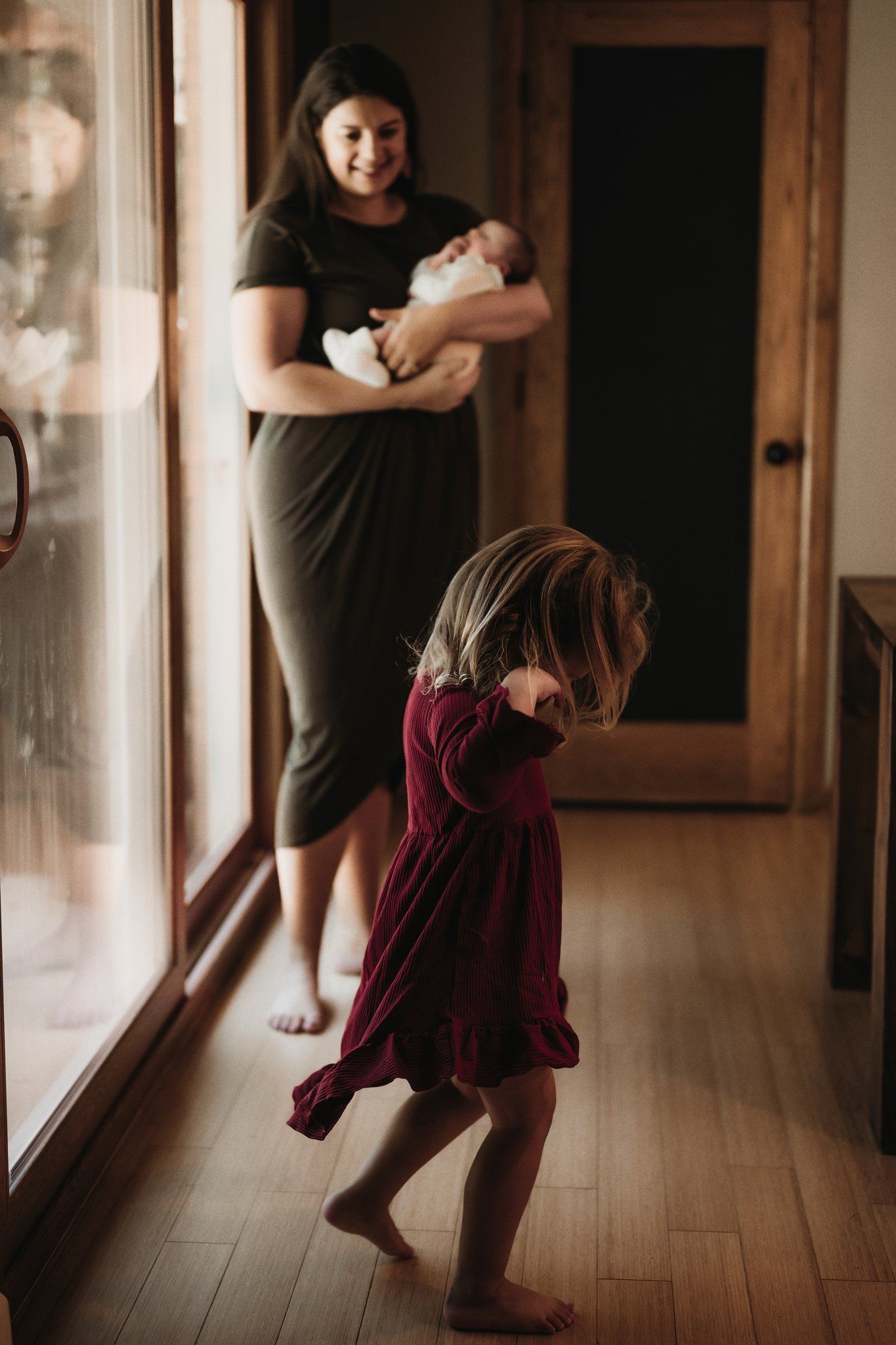 A family moment by large windows as toddler dances while parent holds infant.